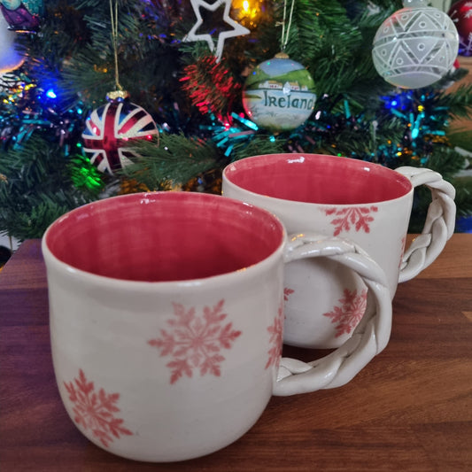 Two mugs with pink snowflake patterns in front of a Christmas tree