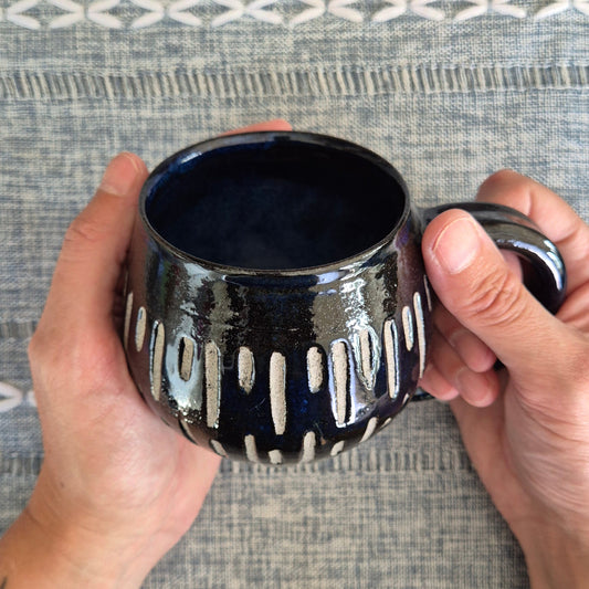 Person holding a dark blue/black ceramic mug with white patterns against a textured gray background
