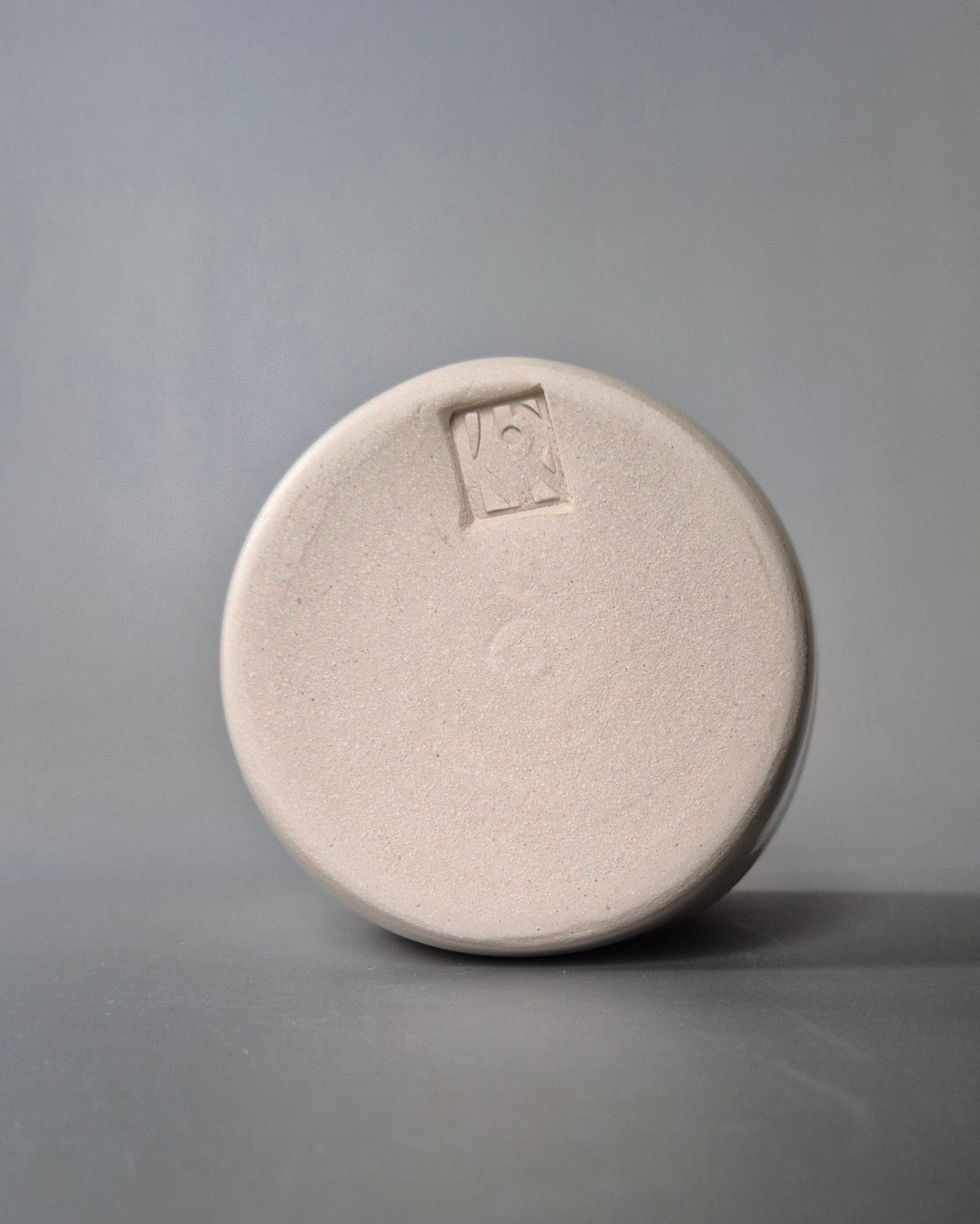 Photo showing the base of a small white stoneware pot with the maker's logo. On a grey background.