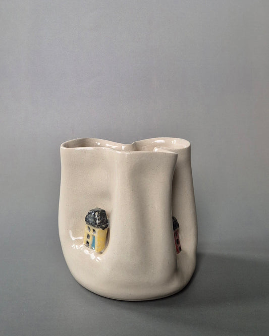 Small white stoneware pot folded to embrace tiny yellow and red cottages. On a grey background