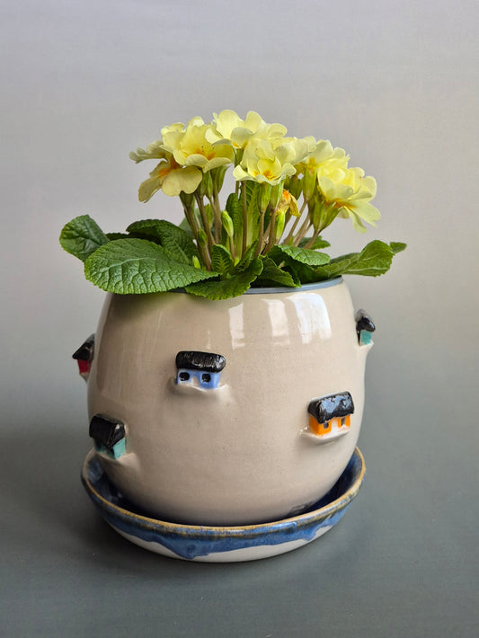 Ceramic planter decorated with colourful tiny houses, with a plant inside. On a blue saucer with a plain background