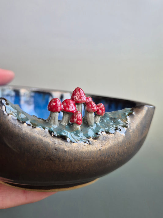 Close up of a small bowl featuring hand sculpted tiny toadstools on the rim. Bronze glazed exterior. The bowl is resting in a person's hand. On a grey background