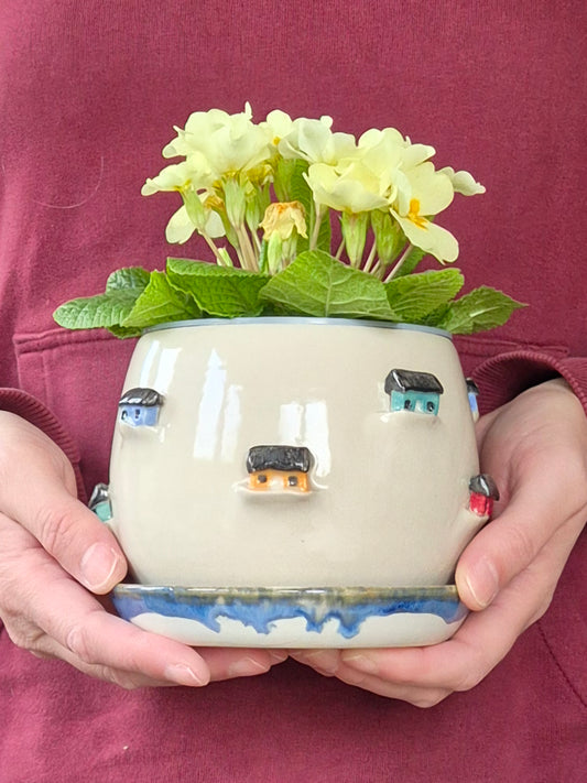 person holding a Ceramic planter decorated with colourful tiny houses. On a blue saucer with a plain background.