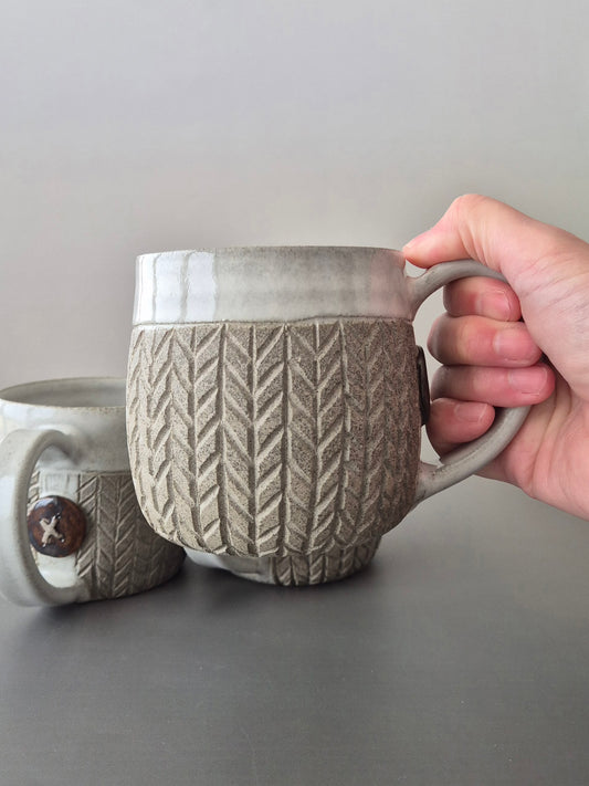 Person holding a textured ceramic mug with another mug in the background on a gray surface.