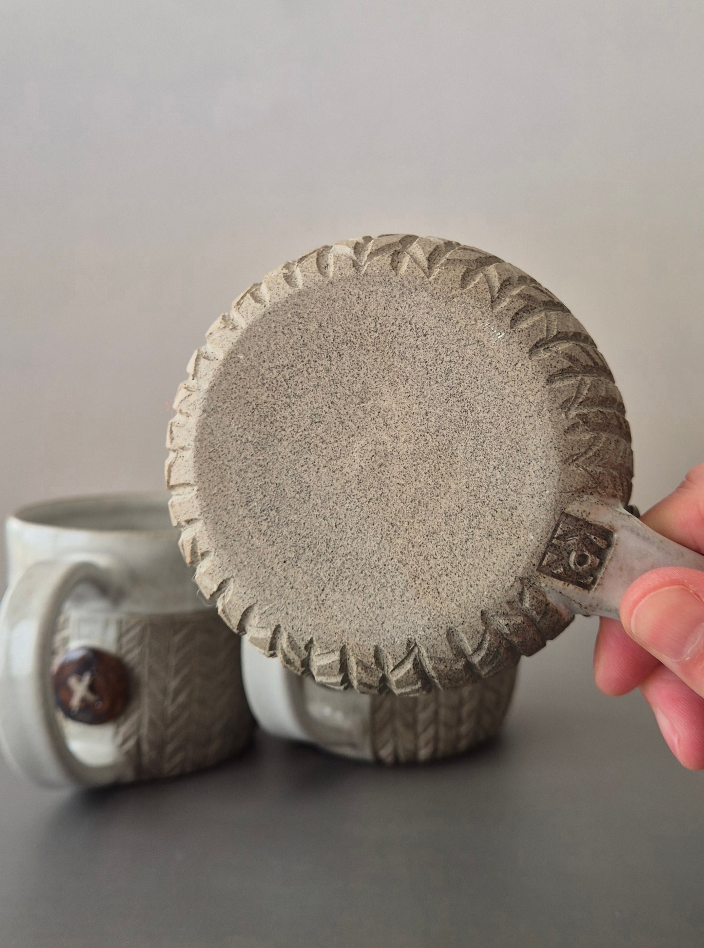 Hand holding a textured ceramic mug to show the base, with other mugs in the background. On a neutral background