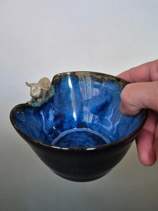 A hand holding a small ceramic bowl featuring a tiny sculpted figure of a highland cow. On a plain grey background.