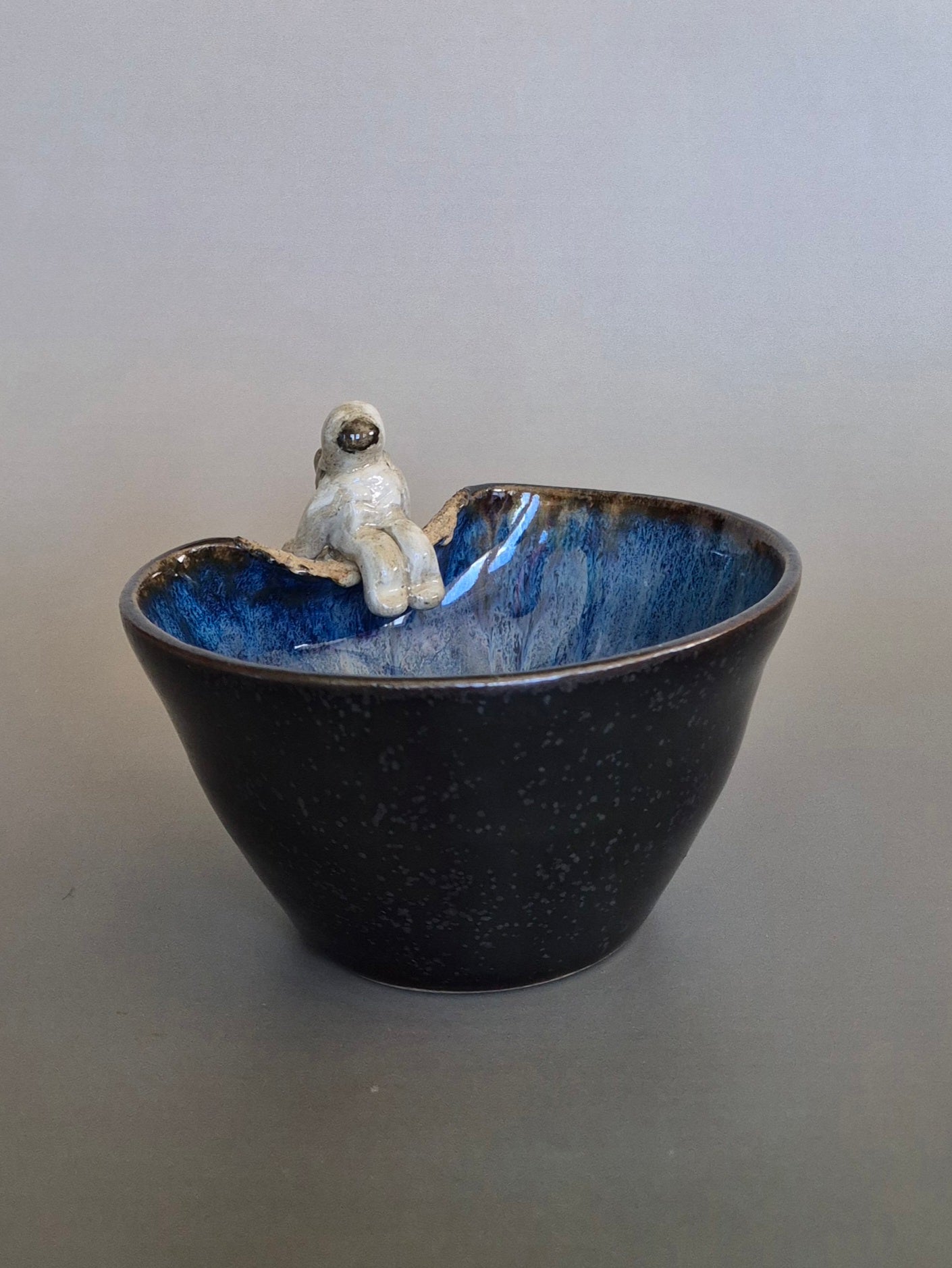 Small figurine of an astronaut sitting on the edge of a bowl. On a plain background