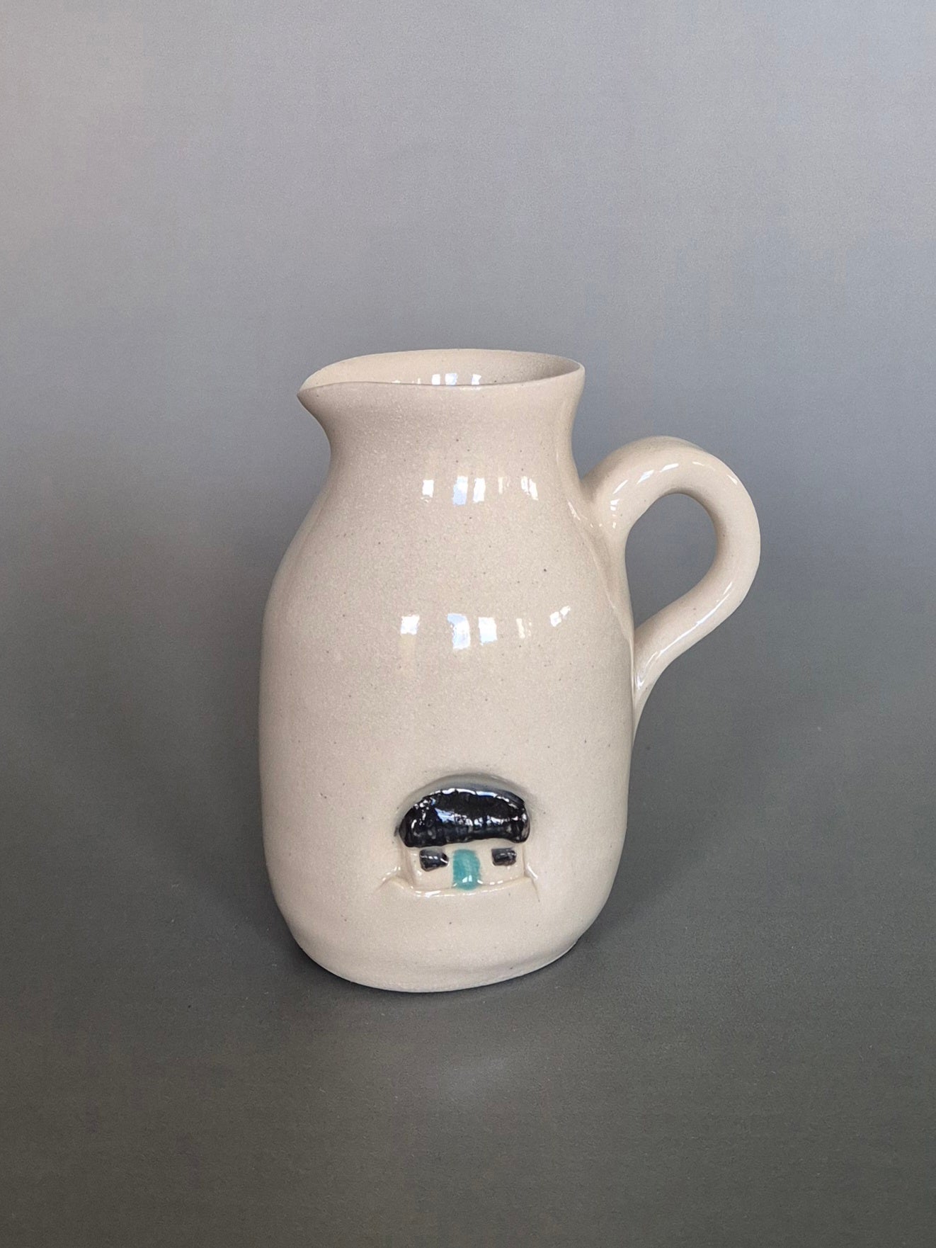 White ceramic pitcher with a small black and blue house design on a gray background