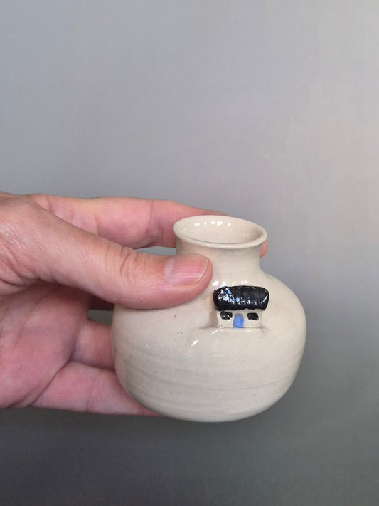 A hand holding a small white ceramic bottle or bud vase. On a plain background
