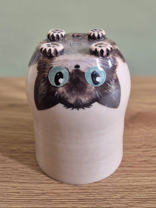A ceramic cat-shaped mug with a glossy finish, featuring a cream-colored body, dark brown and black fur details, and large light blue eyes. It is shown upside down on a wooden surface against a soft green background.