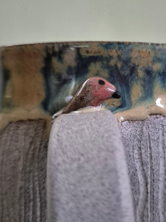 A close-up of a handmade ceramic mug featuring a detailed sculpted bird perched on the rim. The bird has a pinkish-red head, brown speckled body, and a black beak.