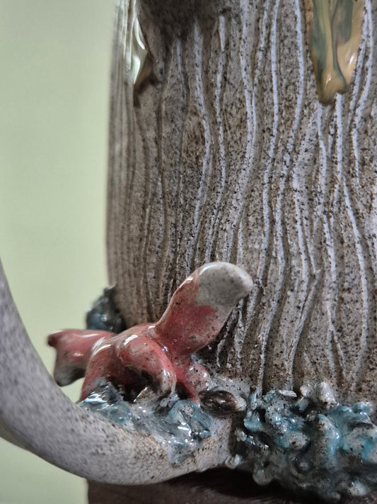 Close-up of a textured ceramic vessel with a sculpted red fox and blue-gray folage detail at the base, set against a soft pale green background.