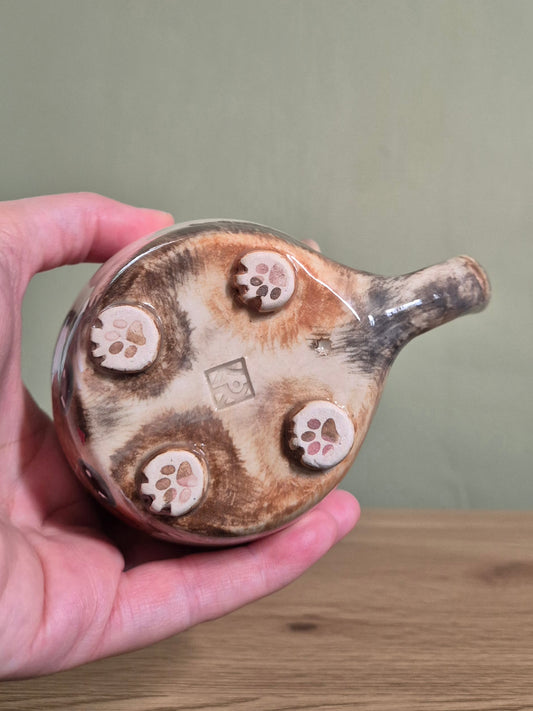A hand holding a small handmade ceramic vessel, showing the bottom with four raised paw-print footprints and a glossy brown glaze on a beige base, against a muted green wall and wooden tabletop.