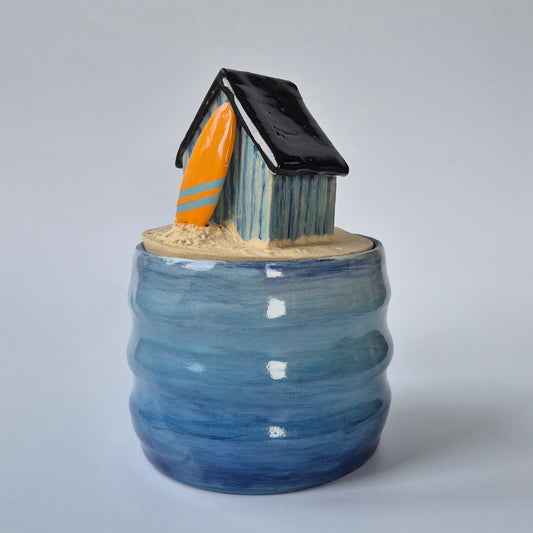 Ceramic jar with a small house and surfboard on top against a gray background