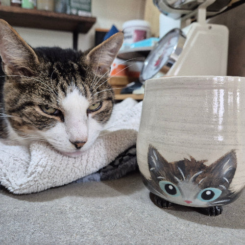 Cat lying next to a ceramic mug with a cat design on a blanket.
