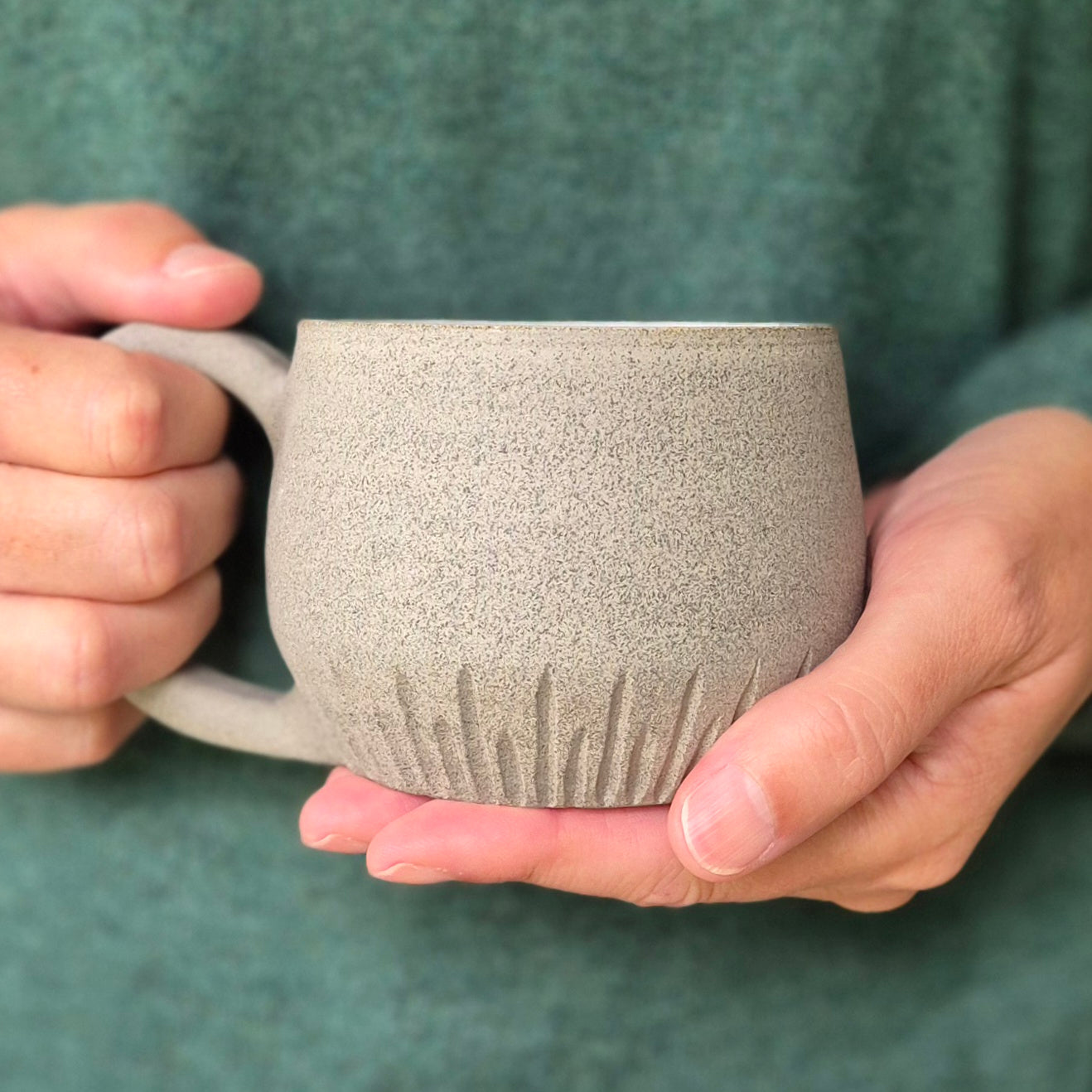 Hands holding a handmade stoneware mug with carved base details and a glossy blue-brown interior glaze; denim blue tablecloth in the background.