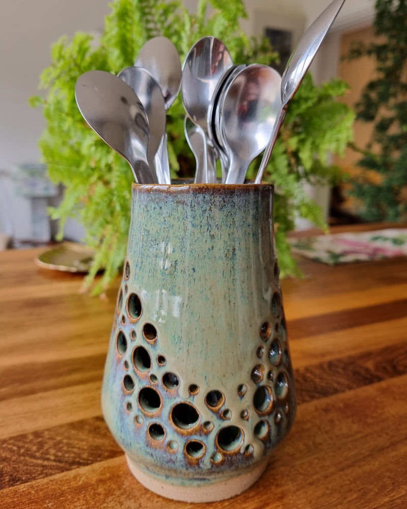 Set of silver spoons with a ceramic holder on a wooden surface with plants in the background