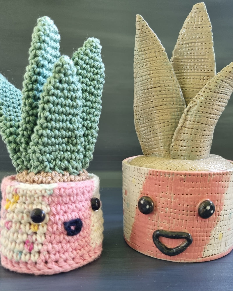 A crochet aloe plant in a pot with a face alongside a ceramic replica. On a dark background