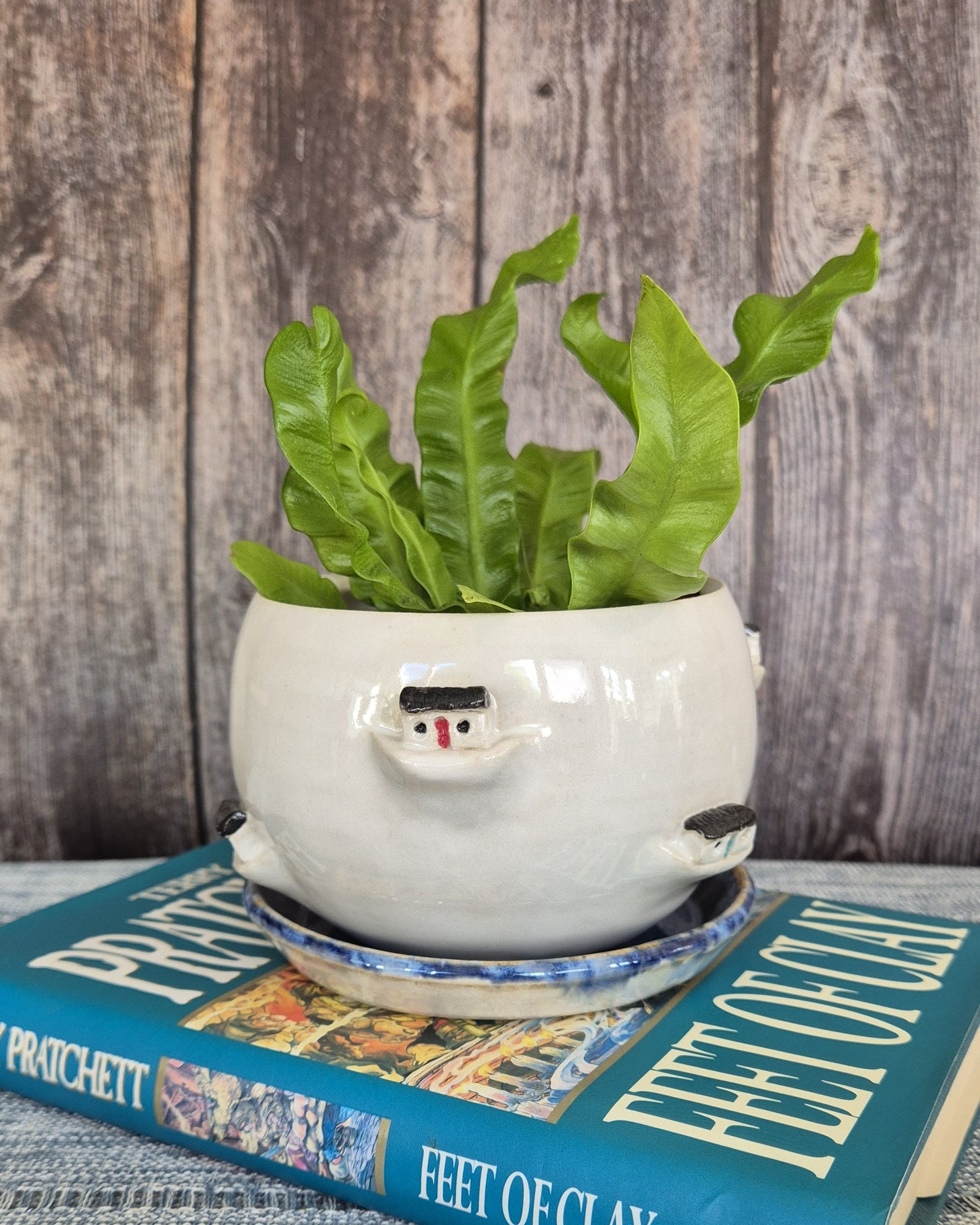 White ceramic planter with sculpted boothy houses on top of a book titled 'Feet of Clay' against a wooden background.