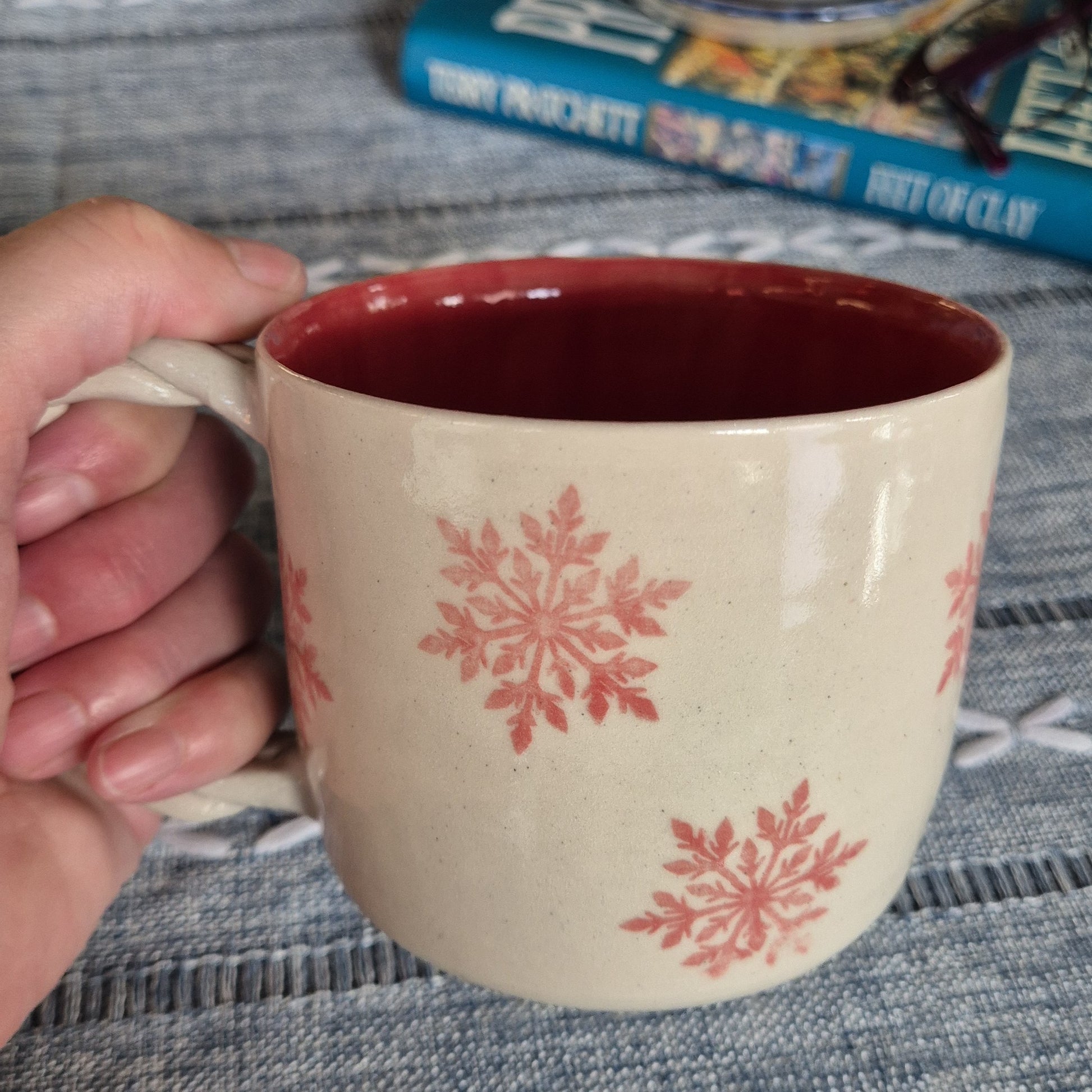 Beige mug with pink snowflake patterns held by a hand on a textured surface.