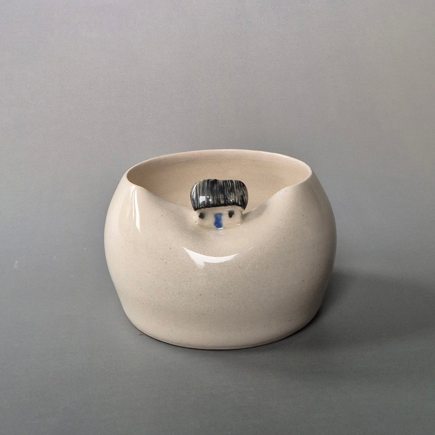 Small cream coloured bud vase featuring a sculptured tiny cottage on the rim. On a grey background