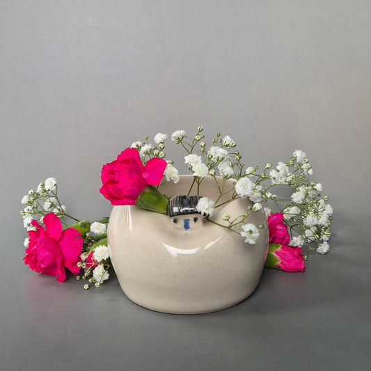 Small cream coloured bud vase featuring a sculptured tiny cottage on the rim. The vase is filled with bright pink carnations and baby's breath with a grey background