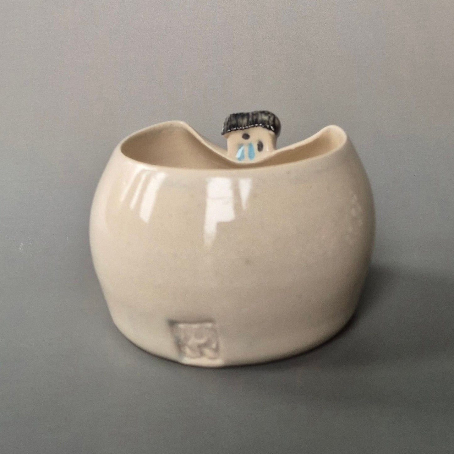 Small cream coloured bud vase featuring a sculptured tiny cottage on the rim. The maker's logo is shown at the back of the vase. On a grey background