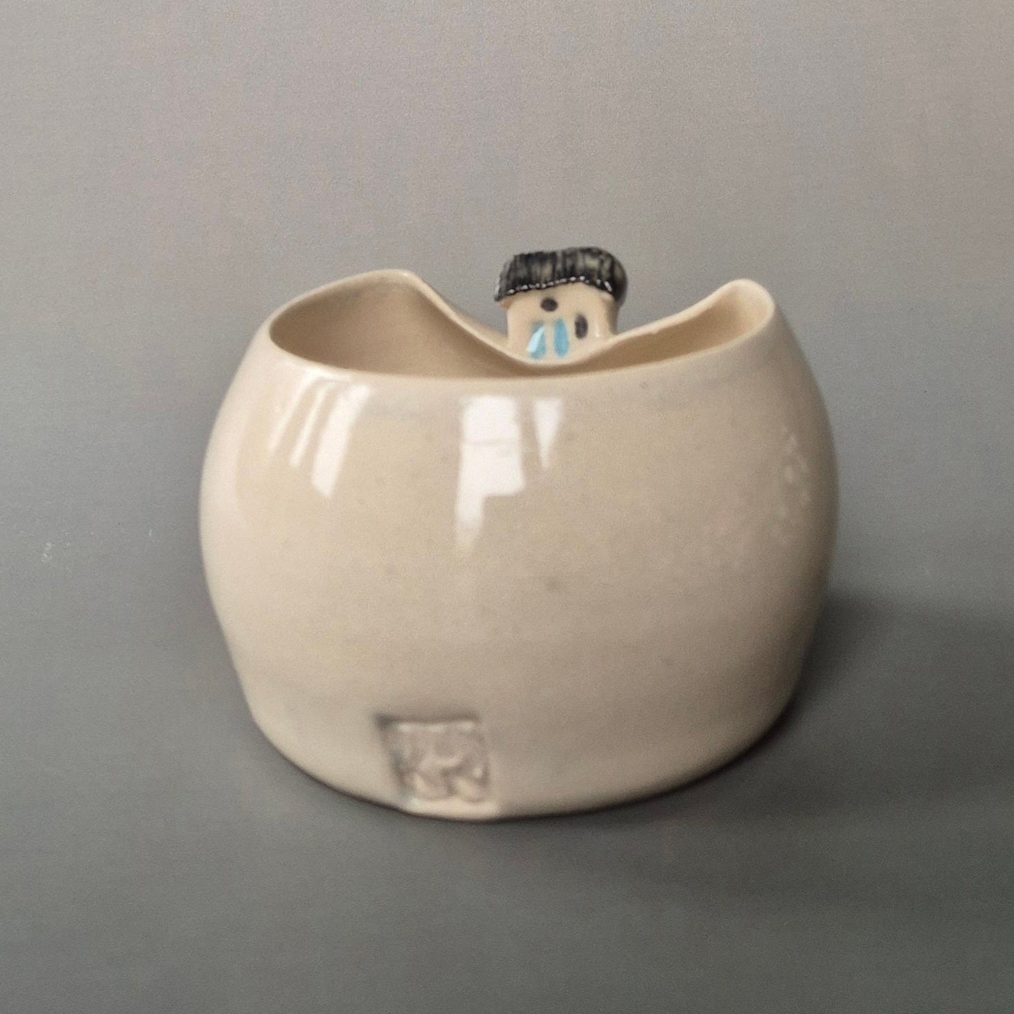 Small cream coloured bud vase featuring a sculptured tiny cottage on the rim. The maker's logo is shown at the back of the vase. On a grey background
