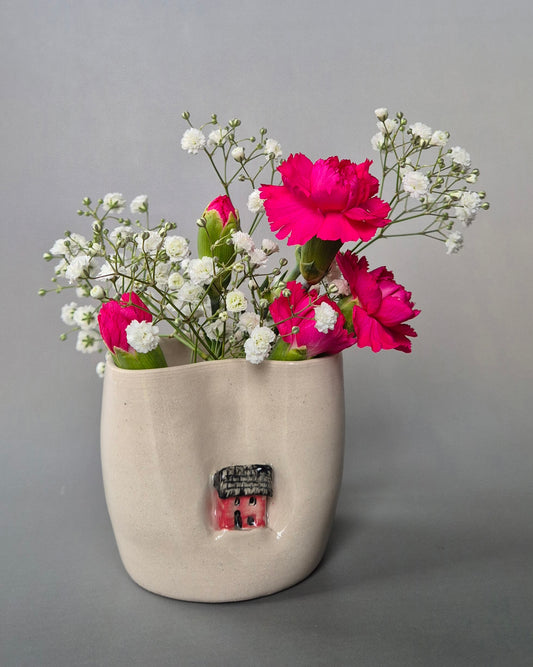 A small white stoneware pot featuring a hand sculpted tiny red house. The pot contains bright pink carnations and white baby's breath flowers. On a grey background.