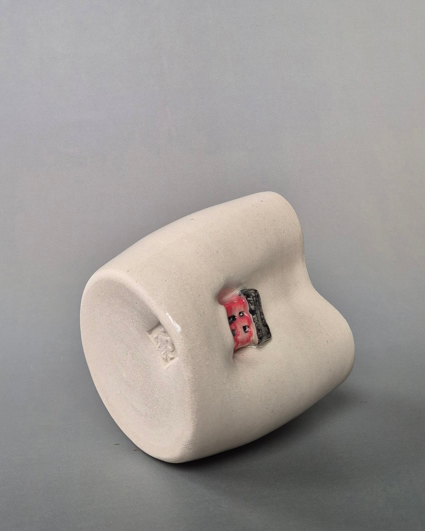 A photo showing the base of a small white heart-shaped stoneware pot featuring a hand sculpted tiny red house. On a grey background.