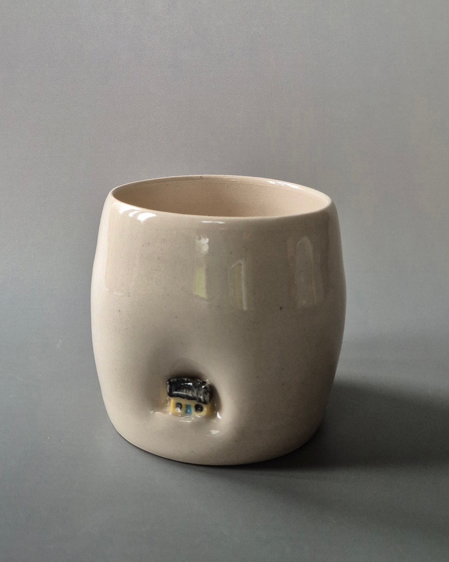 Small white stoneware pot indented with a tiny yellow cottage. On a grey background