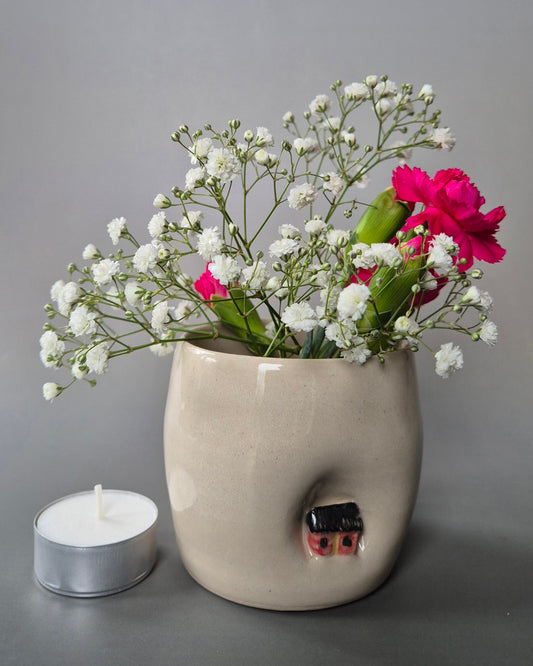 Small white stoneware pot indented to embrace a tiny red cottage. The pot contains bright pink carnations and baby's breath flowers. On a grey background
