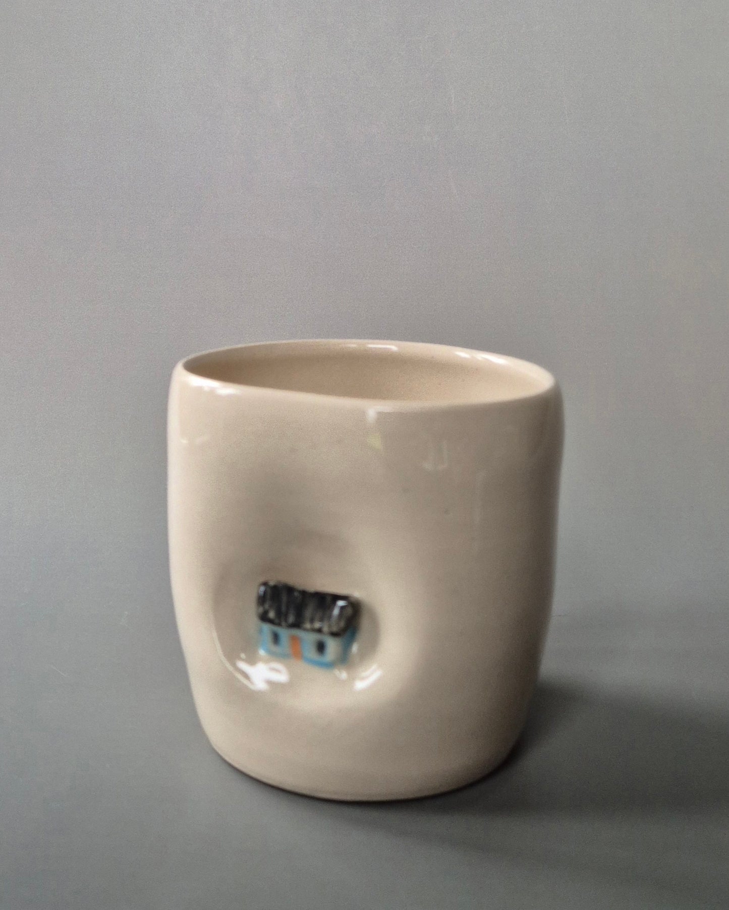 Small white stoneware pot featuring a small hand-sculpted blue cottage embedded in the clay. On a grey background