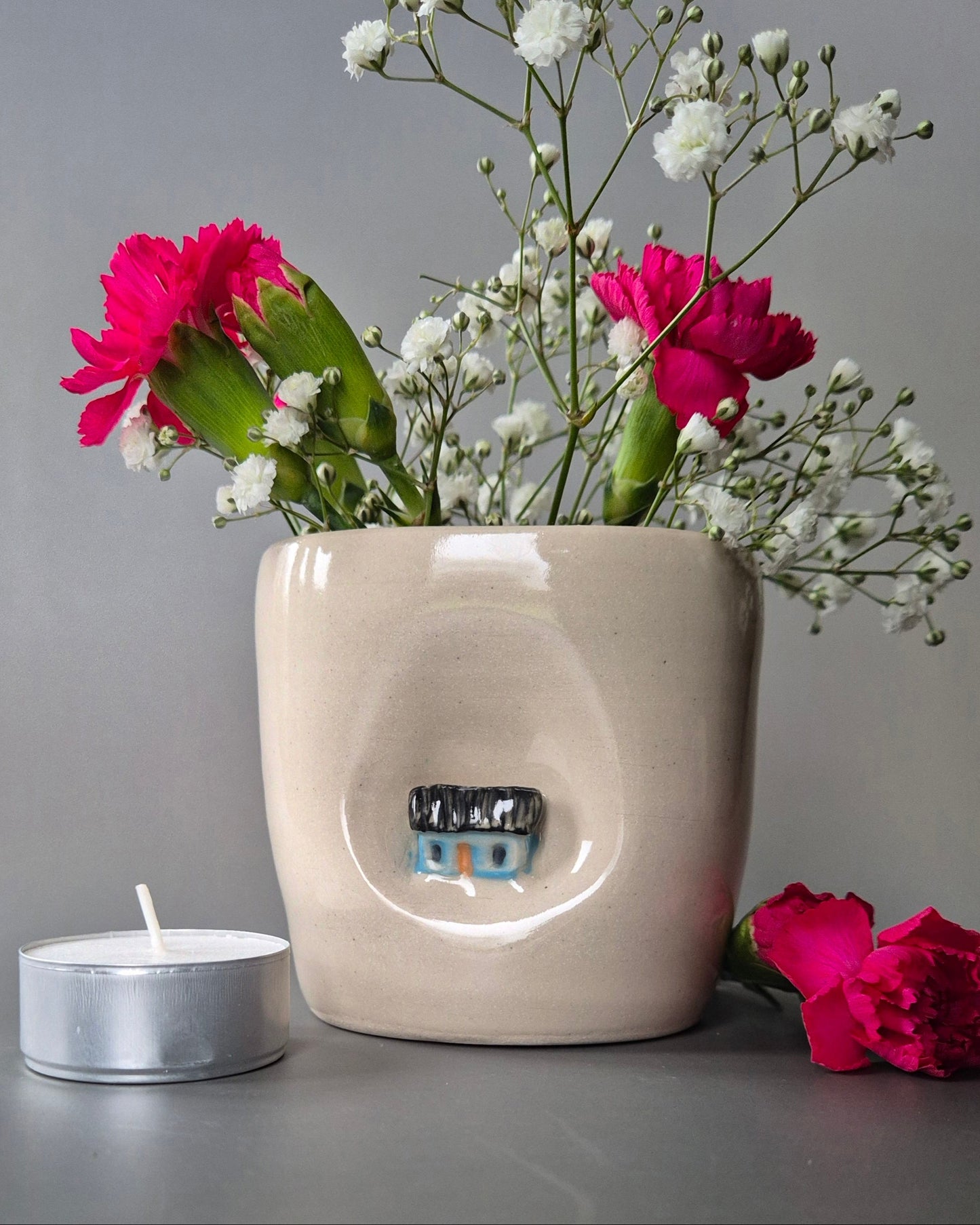 Small white stoneware pot featuring a small hand-sculpted blue cottage embedded in the clay. The pot contains bright pink carnations and baby's breath and is next to a tea-light candle on a grey background