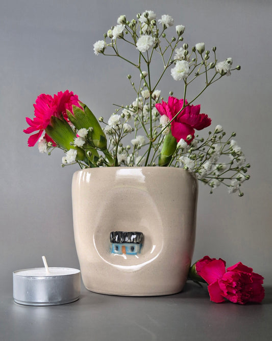 Small white stoneware pot featuring a small hand-sculpted blue cottage embedded in the clay. The pot contains bright pink carnations and baby's breath and is next to a tea-light candle on a grey background
