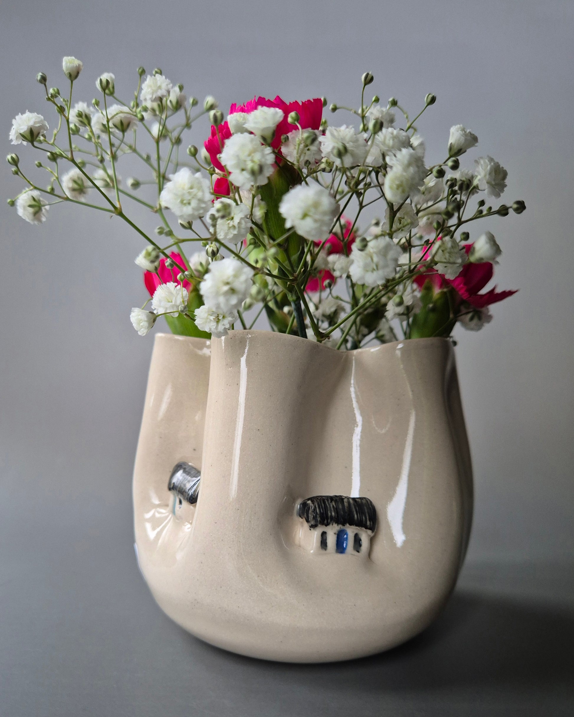 Small white stoneware pot folded to embrace a tiny farm cottages. The pot contains bright pink carnations and baby's breath flowers. On a grey background
