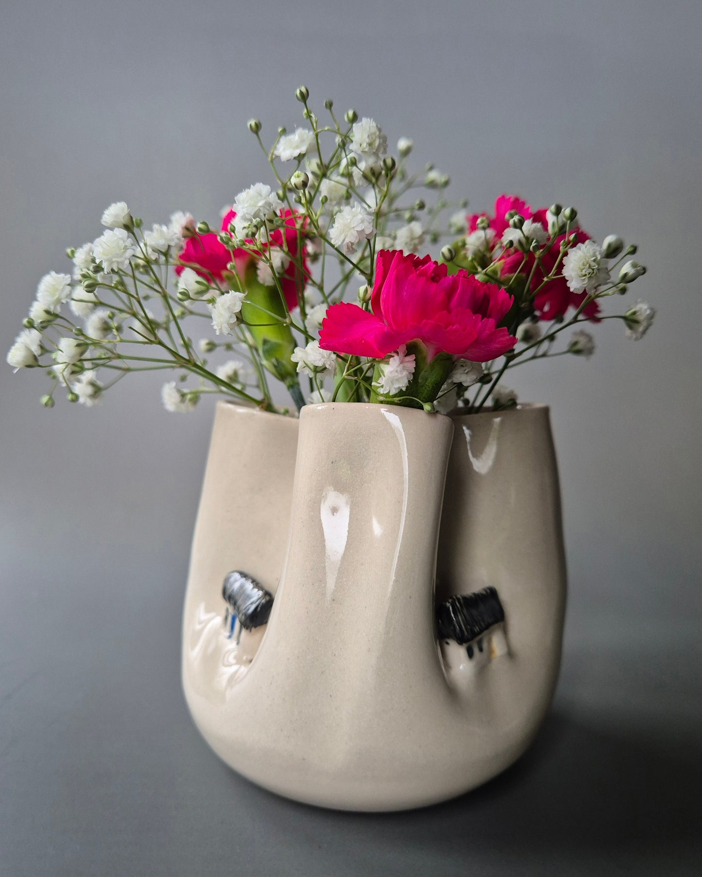 Small white stoneware pot folded to embrace a tiny farm cottages. The pot contains bright pink carnations and baby's breath flowers. On a grey background