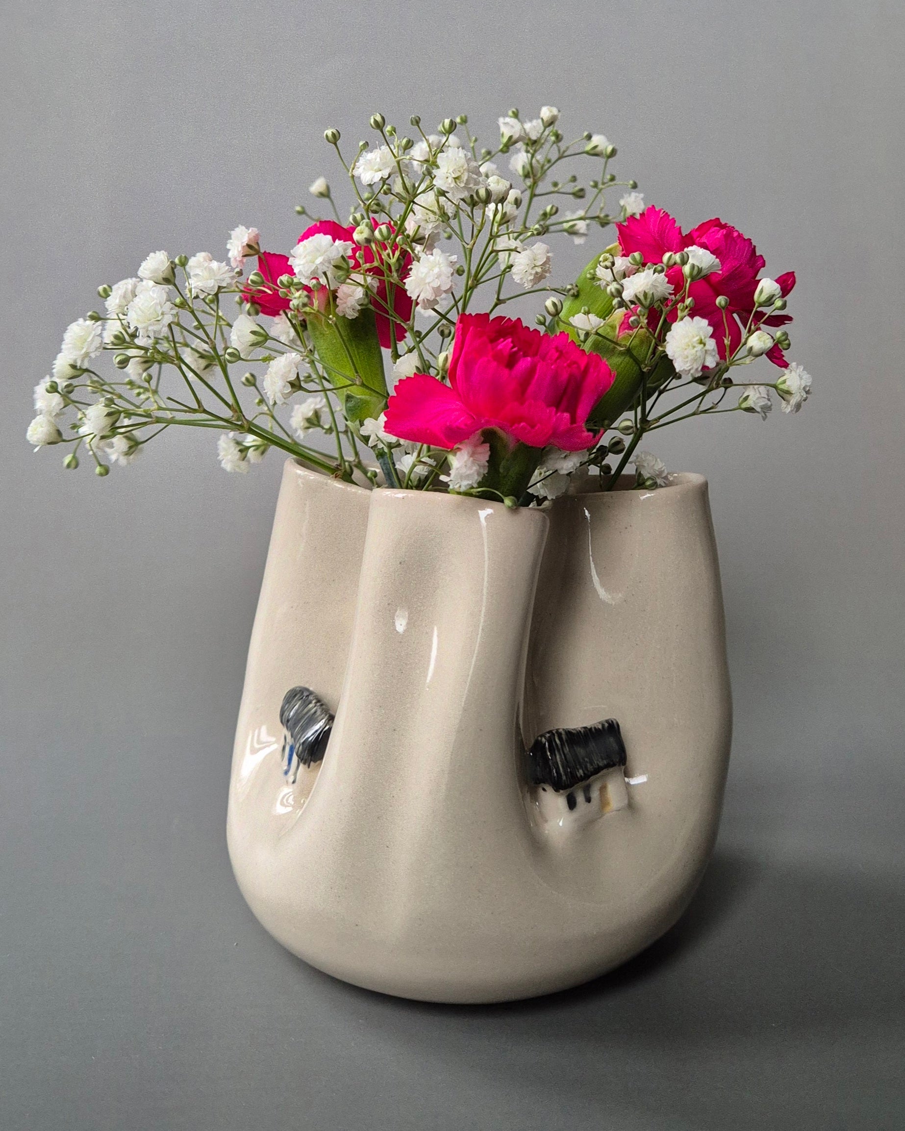 Small white stoneware pot folded to embrace tiny farm cottages. The pot contains bright pink carnations and baby's breath flowers. On a grey background