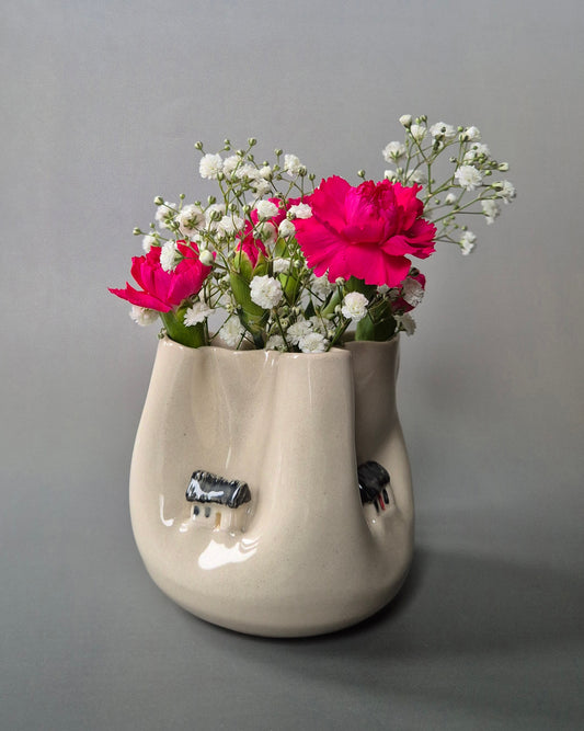 Small white stoneware pot folded to embrace tiny farm cottages. The pot contains bright pink carnations and baby's breath flowers. On a grey background