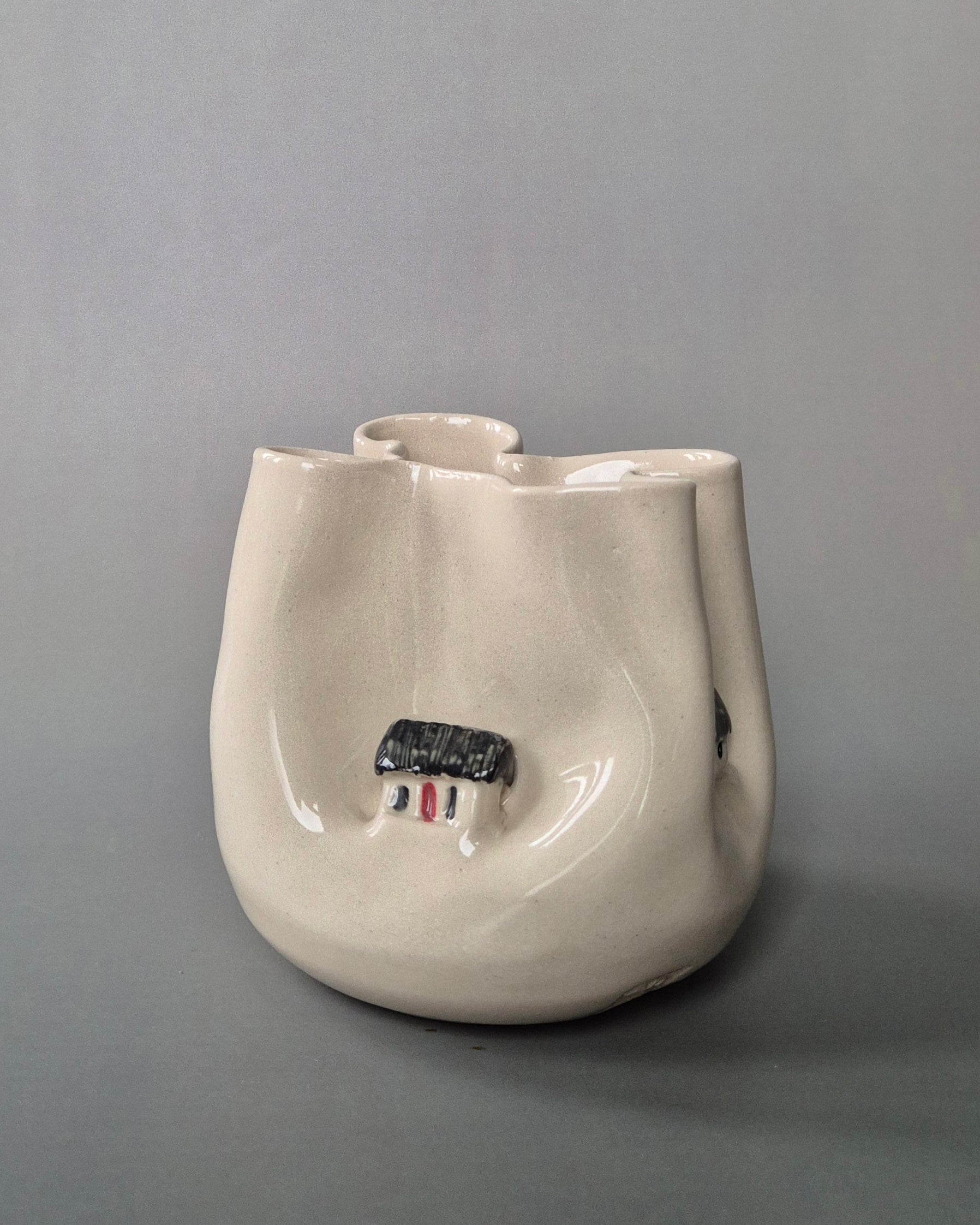 Small white stoneware pot folded to embrace a tiny white cottages. On a grey background