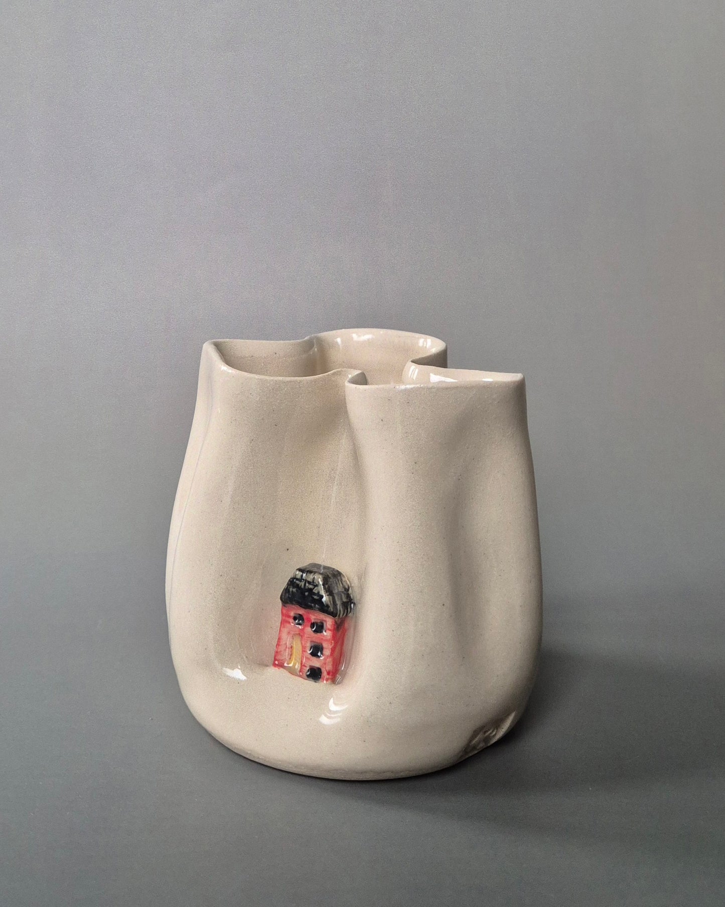 Small white stoneware pot folded to embrace a tiny red cottage. On a grey background