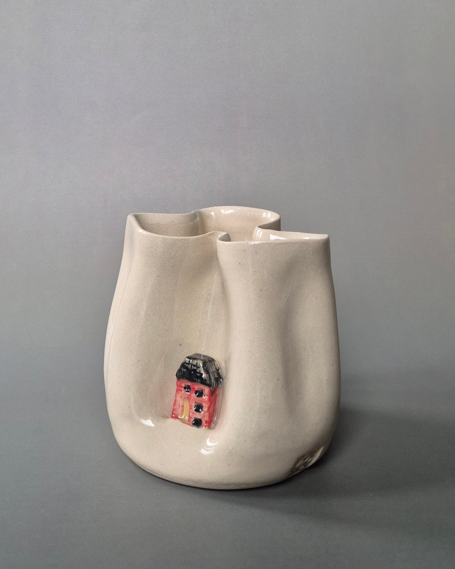 Small white stoneware pot folded to embrace a tiny red cottage. On a grey background