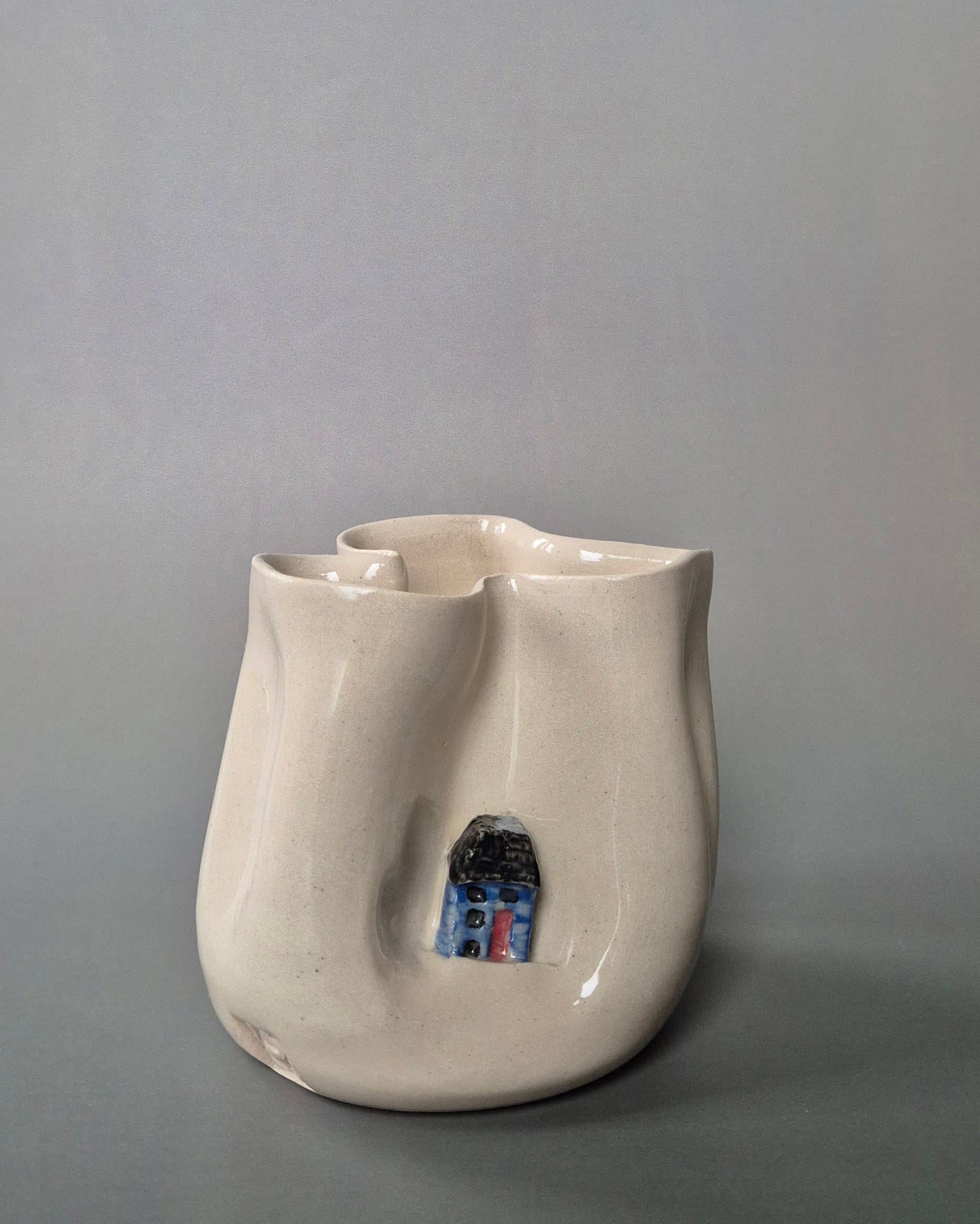 Small white stoneware pot folded to embrace a tiny blue cottage. On a grey background