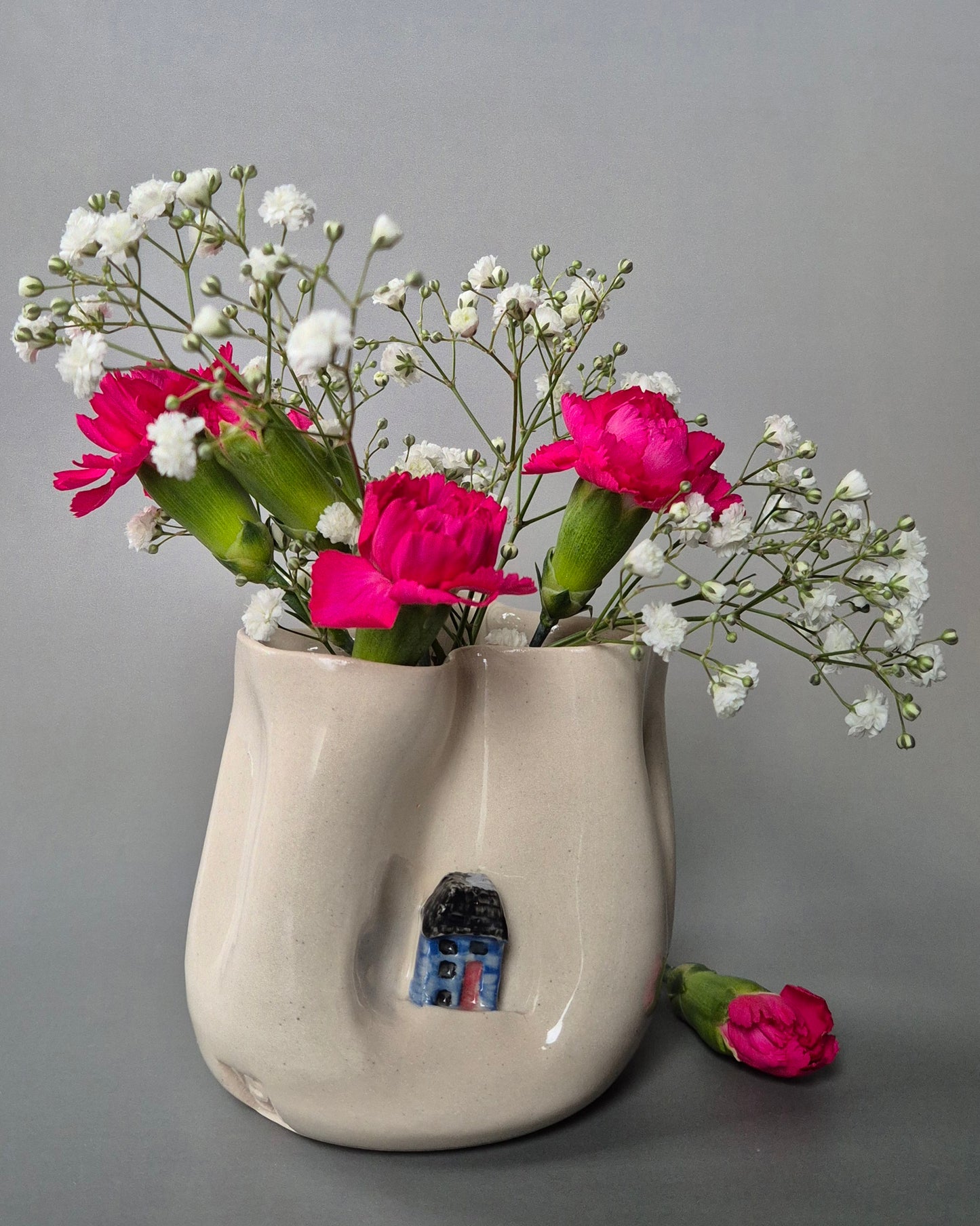 Small white stoneware pot folded to embrace a tiny blue cottage. The pot contains bright pink carnations and baby's breath flowers. On a grey background