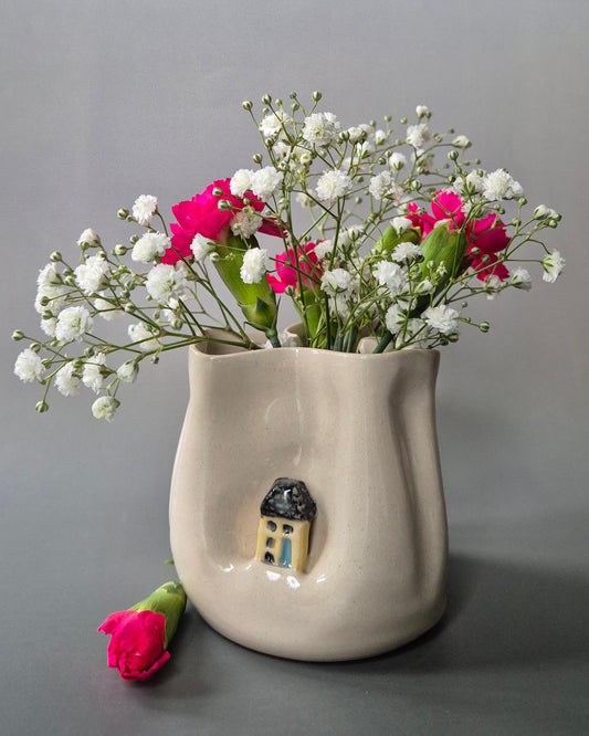 Small white stoneware pot folded to embrace a tiny yellow cottage. The pot contains bright pink carnations and baby's breath flowers. On a grey background