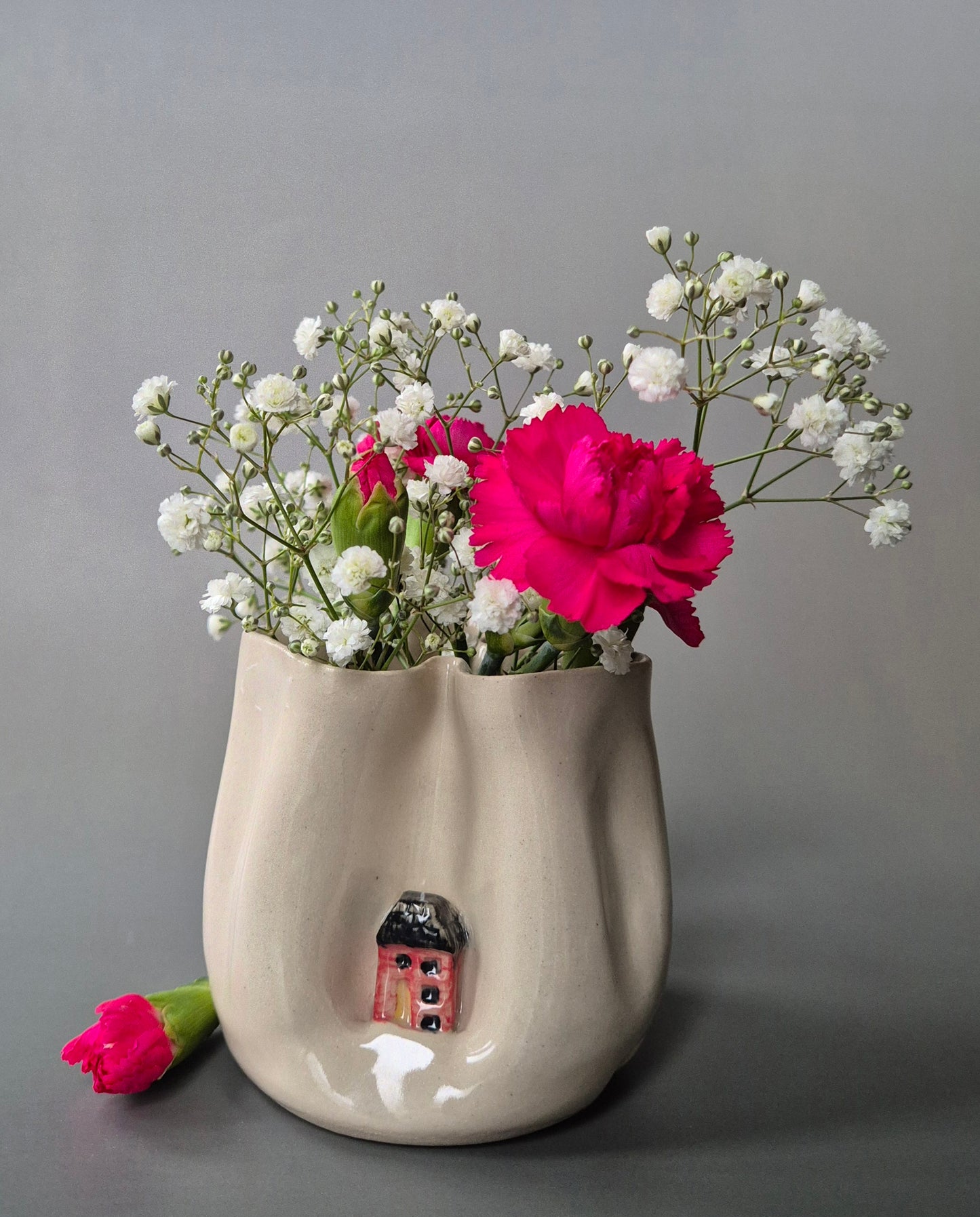 Small white stoneware pot folded to embrace a tiny red cottage. The pot contains bright pink carnations and baby's breath flowers. On a grey background