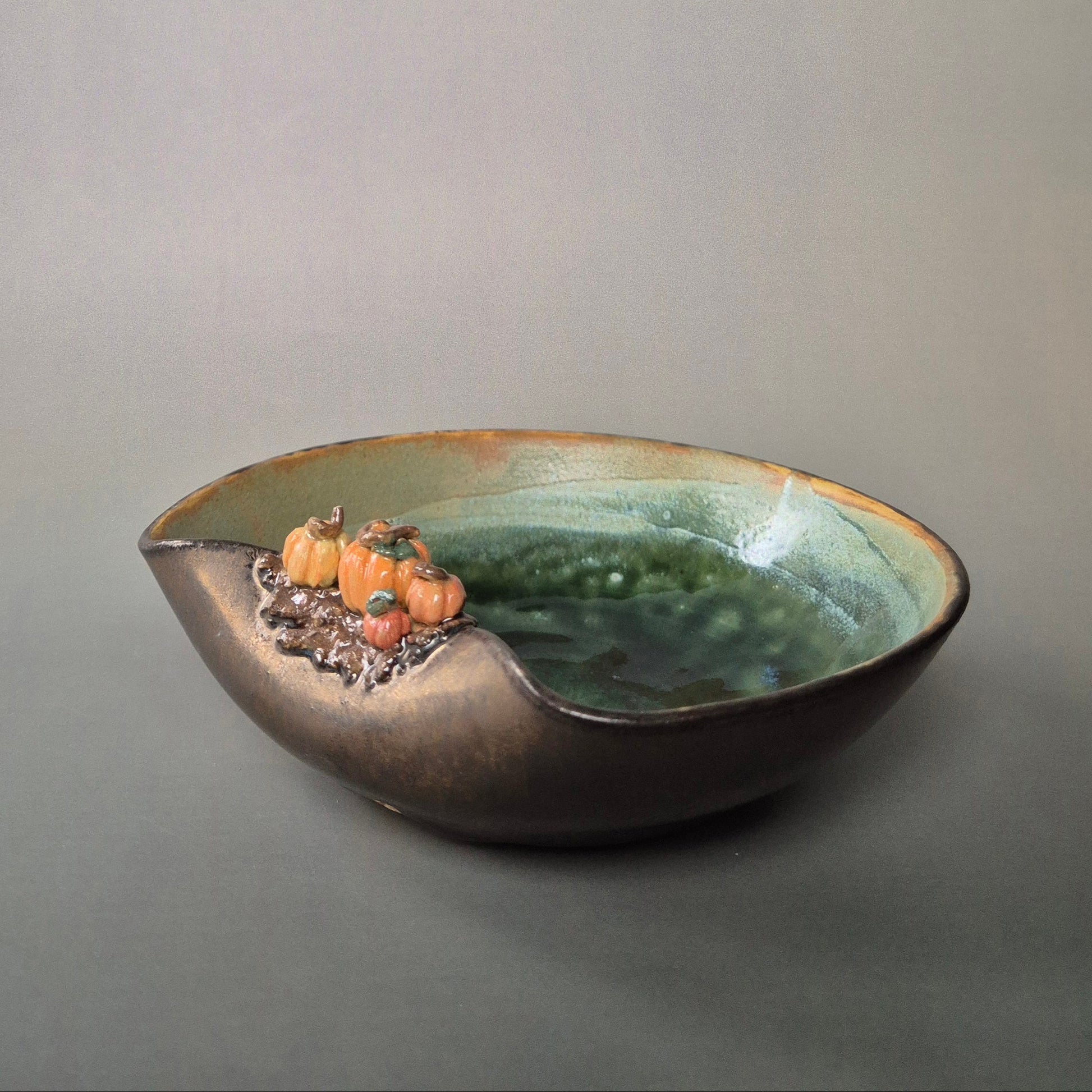 Handmade stoneware trinket dish with green interior and bronze exterior. Decorated with 4 tiny sculpted pumpkins on the bowl edge. There is a grey background.