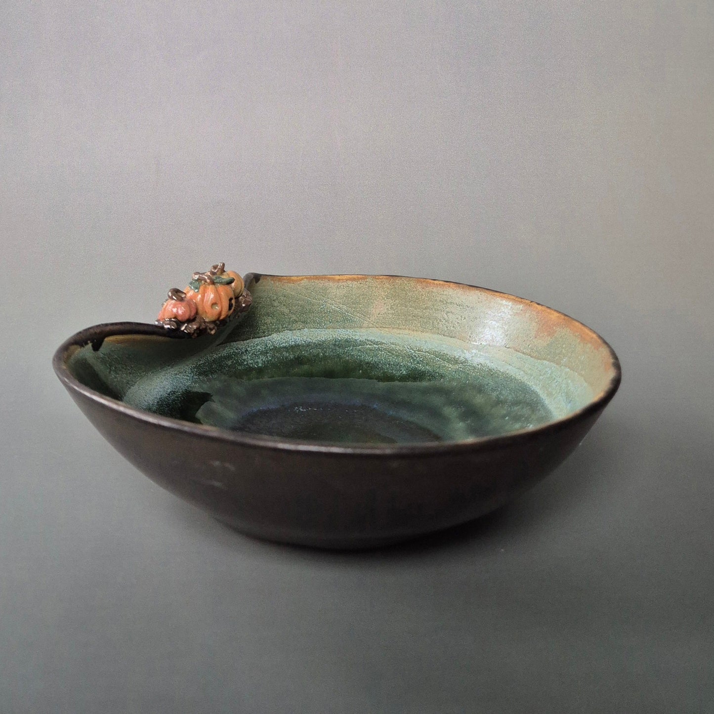 Handmade stoneware trinket dish with green interior and bronze exterior. Decorated with tiny sculpted pumpkins on the bowl edge. There is a grey background.