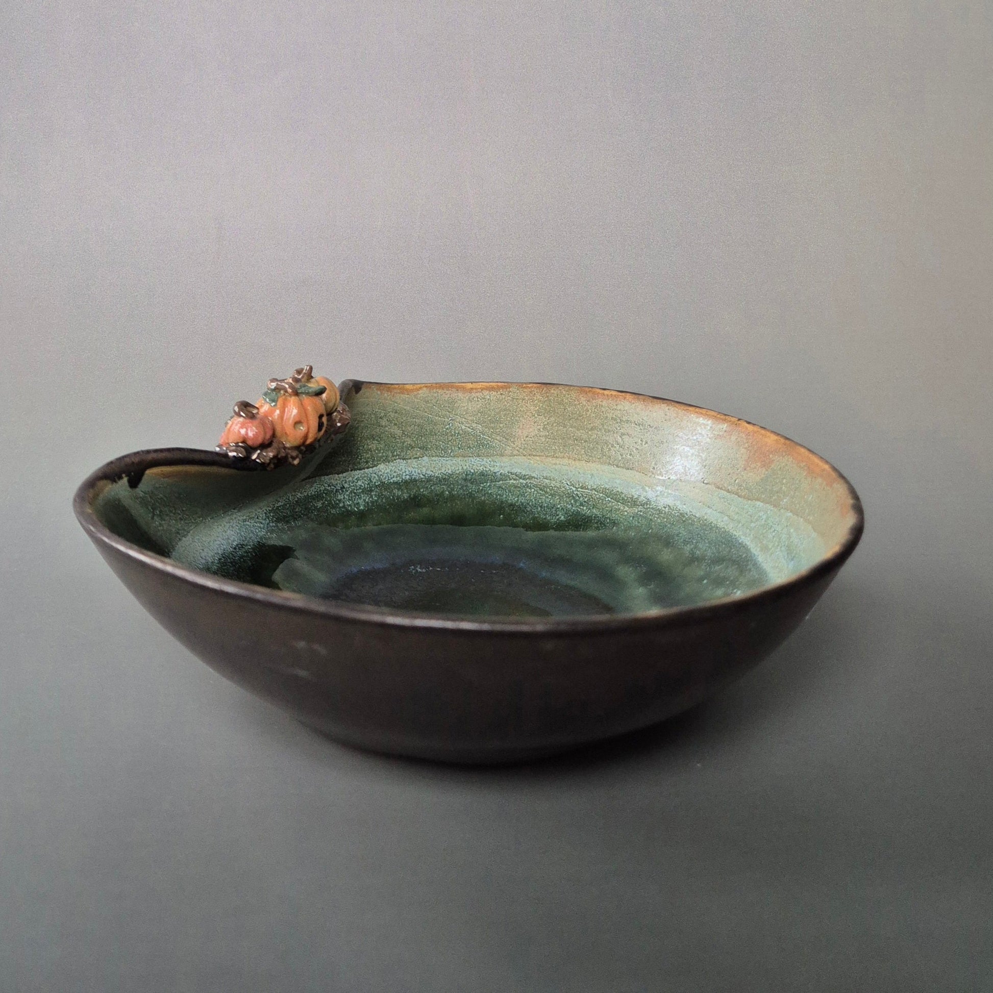 Handmade stoneware trinket dish with green interior and bronze exterior. Decorated with tiny sculpted pumpkins on the bowl edge. There is a grey background.