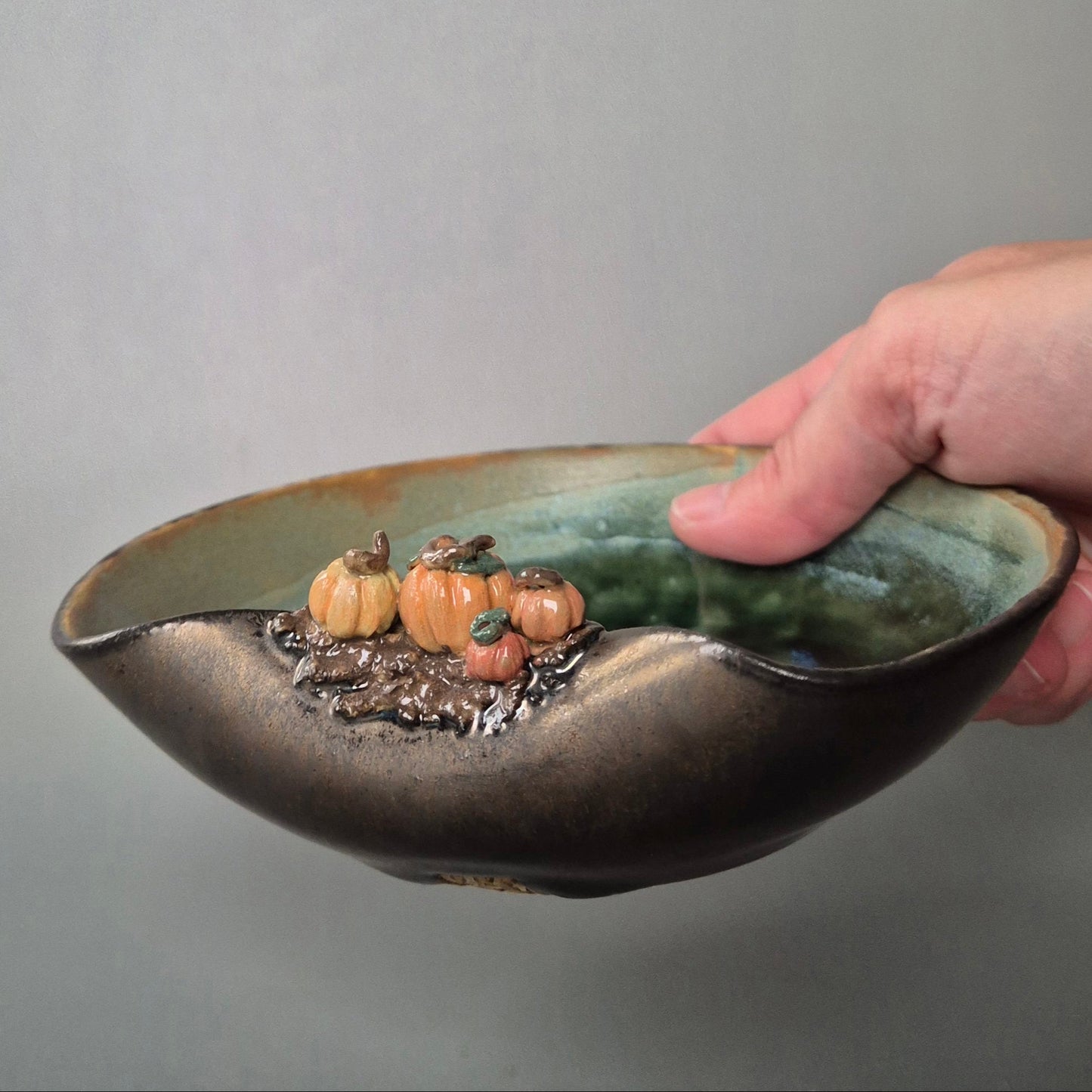 Hand holding a handmade stoneware trinket dish with tiny sculpted pumpkins on the rim, green glazed interior and bronze exterior.